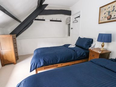 A bedroom with two single beds with blue bedding a wooden chest of drawers a wooden nightstand with a blue lamp and a painting on the wall at Lime Cottage in Burton Bradstock