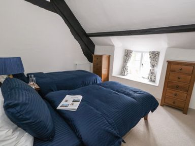 A bedroom with two single beds blue bedding wooden dressers and a window with patterned curtains at Lime Cottage in Burton Bradstock