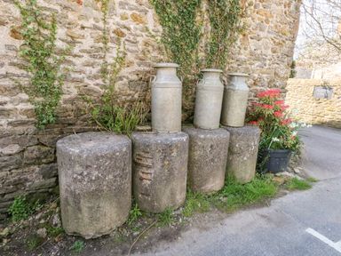 Three metal milk churns on stone bollards next to a stone wall with ivy and flowers at Lime Cottage in Burton Bradstock