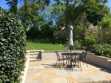 An outdoor patio area with a round table four chairs and a closed umbrella a short set of steps leads to a grassy garden with trees and lattice fencing at Lime Cottage Burton Bradstock