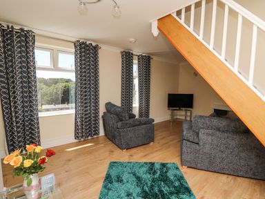 A living room with gray sofas blue patterned curtains a green rug a glass table with flowers and a TV under a staircase at 2 Sunny Hill in Porthgain near St Davids