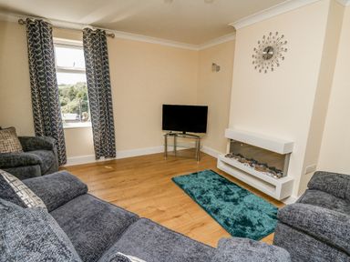 A living room with blue curtains a wall clock a television on a glass stand a blue rug and grey sofas at 2 Sunny Hill in Porthgain near St Davids