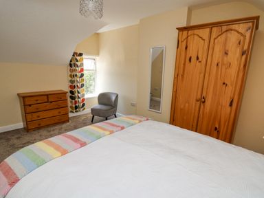 A bedroom with a white bedspread and rainbow blanket a pine wardrobe a chest of drawers a grey chair and patterned curtains at 2 Sunny Hill in Porthgain near St Davids