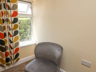 A gray cushioned chair near a window with patterned curtains in a room at 2 Sunny Hill in Porthgain near St Davids