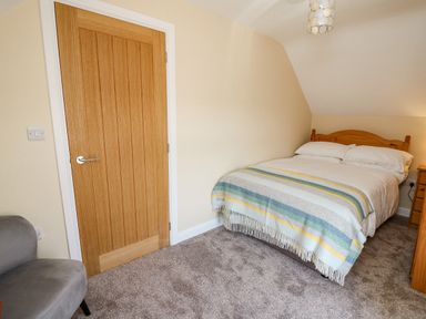 A bedroom with a double bed covered with a striped blanket a wooden door a grey armchair and carpeted floor at 2 Sunny Hill in Porthgain near St Davids