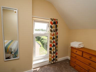 A corner of a bedroom with a window, colorful patterned curtain, wooden chest of drawers with towels, and a wall-mounted mirror at 2 Sunny Hill in Porthgain near St Davids