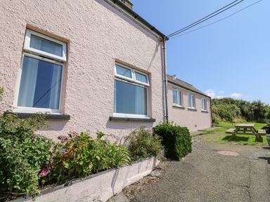 Exterior view of a pink textured wall building with multiple windows and a flower bed next to a gravel path and a wooden picnic table with grass area at 2 Sunny Hill in Porthgain near St Davids