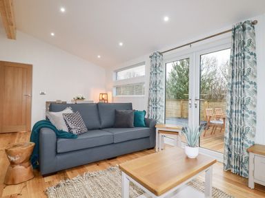 A living room with a gray sofa and patterned curtains next to glass doors showing an outdoor deck at 7 Valley View in Lanreath