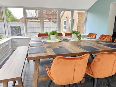 A dining room with a wooden table set with placemats orange chairs and plants by large windows at Sanderson House in Cleator Moor