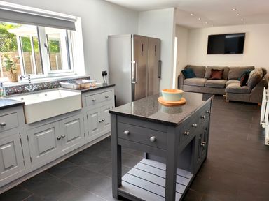 A kitchen with a grey island, white sink under a window, two stainless steel refrigerators and a grey sectional sofa with pillows at Sanderson House in Cleator Moor