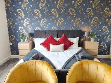 A bedroom with a bed with red and white pillows bedside tables with lamps and plants and two yellow chairs at Sanderson House in Cleator Moor