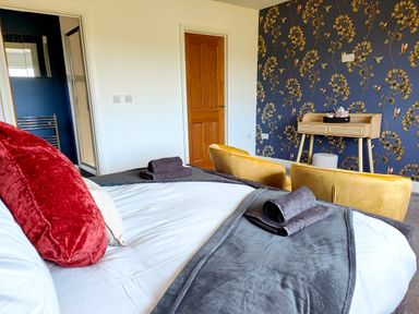 A bedroom with a bed with pillows and towels a wooden door two yellow chairs a wooden desk and floral wallpaper at Sanderson House in Cleator Moor