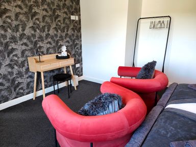 A room with two red armchairs with pillows a small wooden desk with a stool a clothing rack with hangers and patterned wallpaper at Sanderson House in Cleator Moor