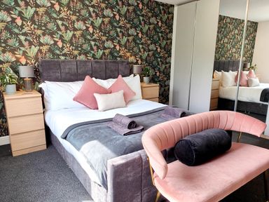 A bedroom with a double bed with pillows and folded towels bedside tables with lamps floral wallpaper and a pink upholstered bench at Sanderson House in Cleator Moor