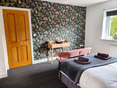 A bedroom with a bed folded towels a pink bench a wooden desk floral wallpaper and a window at Sanderson House in Cleator Moor
