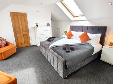 A bedroom with a gray upholstered bed with orange pillows a white dresser next to a wooden door and an orange chair at Sanderson House in Cleator Moor