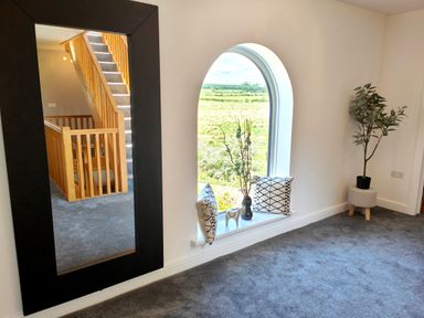 A room with a large black-framed mirror reflecting a wooden staircase and gray carpet a window seat with cushions a vase and a wooden horse and a plant on a small stool at Sanderson House in Cleator Moor