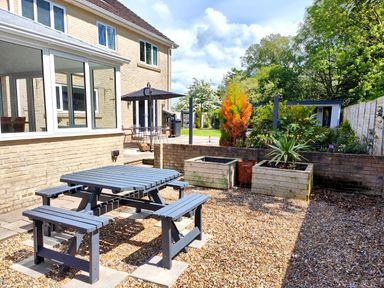 An outdoor garden area with a wooden picnic table and benches next to a house with large windows and a patio with an umbrella and chairs at Sanderson House in Cleator Moor