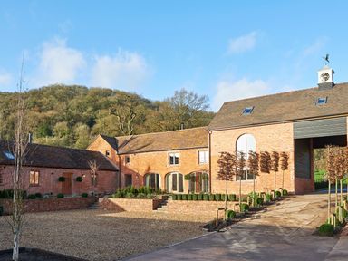 A brick building with multiple windows and a clock tower on the roof surrounded by gravel and small trees at Fairoaks Barns in Hollybush near Ledbury