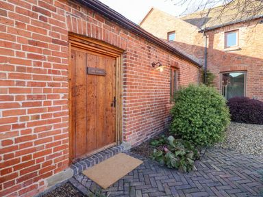 An exterior view of a red brick building with a wooden door labeled The Stables bushes and plant beds nearby at Fairoaks Barns in Hollybush near Ledbury