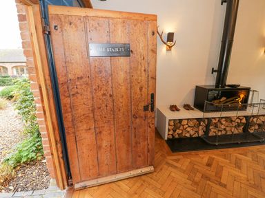 A wooden door with a sign that reads The Stables opening into a room with a fireplace with wood storage underneath and shoes on a bench at Fairoaks Barns Hollybush near Ledbury