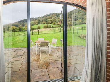 A patio with a table and four chairs outside glass doors overlooking a grassy area with hills and trees at Fairoaks Barns Hollybush near Ledbury