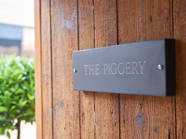 A metal sign with the text THE PIGGERY on a wooden surface with greenery in the background at Fairoaks Barns in Hollybush near Ledbury