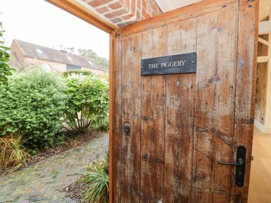 An open wooden door with a sign that reads The Piggery showing a garden with bushes and a brick path at Fairoaks Barns in Hollybush near Ledbury