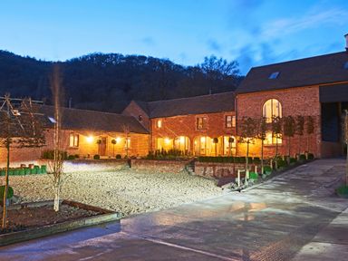 A brick barn building with lit windows and a gravel courtyard surrounded by trees at Fairoaks Barns in Hollybush near Ledbury