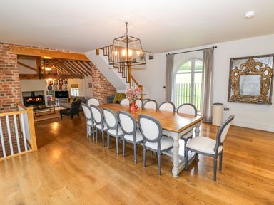 A dining room with a long wooden table and twelve chairs a bouquet of flowers a large window stairs and a living area with a fireplace at Fairoaks Barns in Hollybush near Ledbury