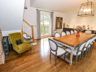 A dining room with a wooden table and twelve chairs a green armchair wooden floor and a window at Fairoaks Barns in Hollybush near Ledbury