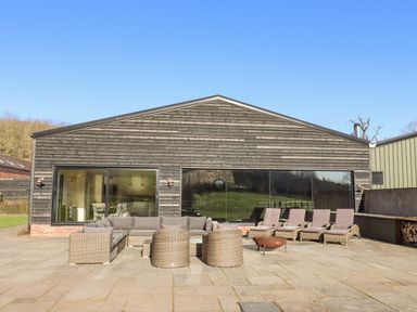 An outdoor patio with wicker chairs sun loungers and a fire pit in front of a wooden building at Fairoaks Barns in Hollybush near Ledbury