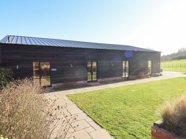 A black barn style building with multiple glass doors and a paved path surrounded by grass at Fairoaks Barns in Hollybush near Ledbury