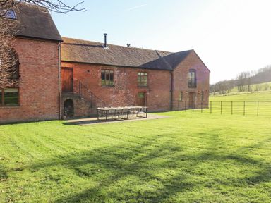 An exterior view of a brick building with a tiled roof and outdoor dining table on a patio surrounded by green grass at Fairoaks Barns Hollybush near Ledbury
