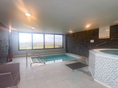An indoor pool area with a small pool a hot tub a wicker chair and large windows overlooking fields at Mallard Cottage in Penrith