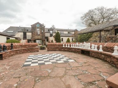 An outdoor courtyard with a large chessboard on the ground and oversized chess pieces on stone benches at Mallard Cottage in Penrith
