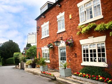 A brick building with white framed windows flowers and plants outside and a hanging sign on a street at No Deer in Ashbourne