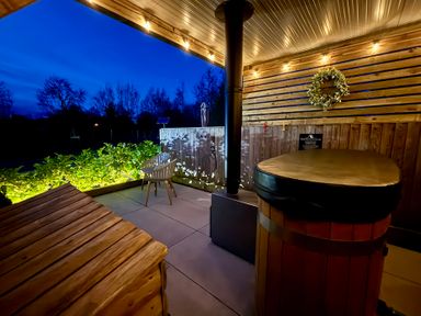 An outdoor patio with a hot tub a wooden bench a chair string lights and plants at No Deer in Ashbourne