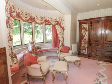 A sitting room with two armchairs, a matching footstool, a curved bench seating by a bay window, floral curtains, and a large wooden wardrobe at Highcliffe Manor in Flamborough