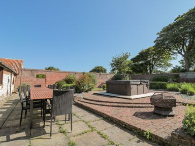 An outdoor patio area with a long wooden table and black chairs a hot tub on a raised brick platform and a fire pit surrounded by a brick wall and trees at Highcliffe Manor in Flamborough