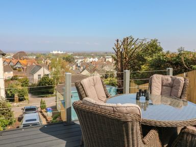 An outdoor seating area with a glass table and wicker chairs overlooking houses and trees at The Outlook in Minehead