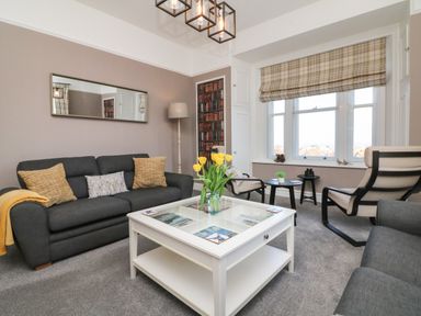 A living room with a dark gray sofa, armchairs, a white coffee table with yellow flowers, a floor lamp, window with plaid blind and small side tables at The Outlook in Minehead