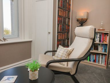 A reading corner with a beige armchair and plaid pillow next to a black round table with a small potted plant and bookshelves at The Outlook in Minehead