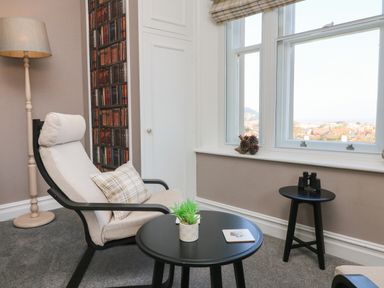 A corner of a room with a beige armchair next to a floor lamp and two black round tables one with a potted plant and one with binoculars by a window at The Outlook in Minehead