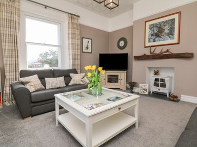 A living room with a gray sofa and cushions a white coffee table with yellow flowers a television on a cabinet and a fireplace at The Outlook in Minehead
