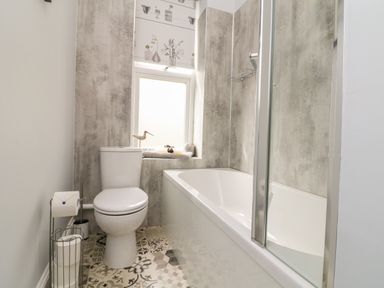 A bathroom with a toilet a bathtub with glass shower doors patterned floor tiles and a window with a patterned blind at The Outlook in Minehead
