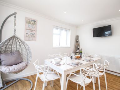 A dining room with a white table and chairs set for a meal a hanging egg chair a wall-mounted television and a corner shelf at The White House in Weymouth