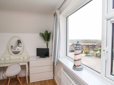 A bedroom corner with a white dressing table and chair a chest of drawers with a small tv and a plant and a window with a lighthouse decoration on the sill at The White House in Weymouth