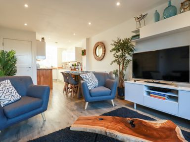A living area with gray armchairs a wooden coffee table a flat screen tv and a dining table with chairs at Chy Fistral in Newquay