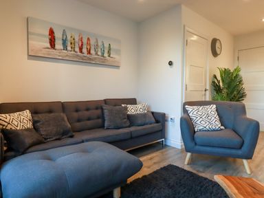 A living room with a gray sofa armchair ottoman pillows wall art clock and plant at Chy Fistral in Newquay